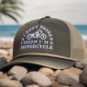 May include: A green and brown trucker hat with a white embroidered design that says "I don't snore I dream I'm a motorcycle" with a motorcycle graphic.