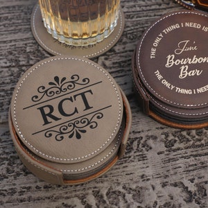 May include: A set of personalized leather coasters in a holder. One coaster features the initials "RCT" with decorative flourishes. Another set of coasters is inscribed with "June's Bourbon Bar" and the phrase "The only thing I need is whisky."
