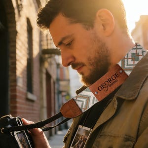 May include: A camera strap with a geometric pattern in teal, white, and red, and a brown leather patch with the name "GEORGE Havens". The strap is attached to a black DSLR camera. A small photo hangs from the strap.