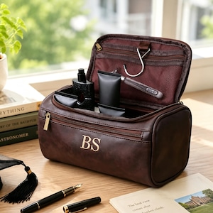May include: A dark brown leather toiletry bag with the gold embroidered initials BS on the front. The bag is open, revealing a dark interior with a metal hanging hook, mesh pockets, and several black toiletry bottles stored inside the main compartment.