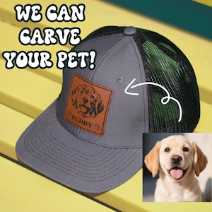 May include: A gray and black trucker hat with a brown leather patch featuring a dog's head and the name "RUDDY". The hat has a mesh back and a curved brim. The text "WE CAN CARVE YOUR PET!" is visible in the background.