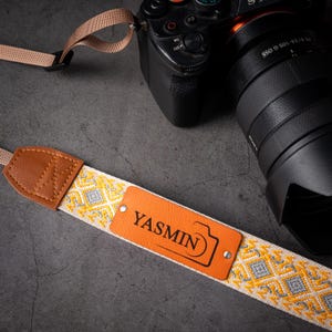 May include: Camera strap with a woven pattern in white, yellow, and gray. The strap has brown leather accents and a brown leather tag that reads "YASMIN" with a camera icon. A black camera with a lens is in the background.