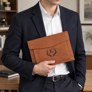 May include: A tan leather document portfolio featuring a flap closure and a centred black monogram of the letter K flanked by two deer antlers. The portfolio has a smooth, matte finish and a structured rectangular shape with clean stitching.
