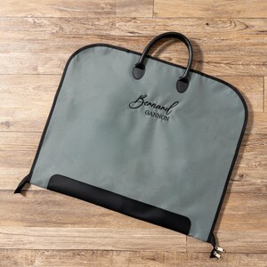 Puede incluir: Una funda de ropa gris con ribetes y asa negros. La bolsa tiene el nombre "Bernard Gannon" en escritura cursiva. Está sobre una superficie de madera.