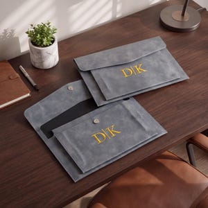 May include: Two grey faux leather document envelopes with a flap closure and a silver snap button. Each envelope features the gold embroidered monogram "D|K" on the front. One envelope is closed, and the other is open, revealing a black interior.