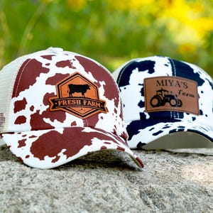 Pode incluir: Dois bonés de caminhoneiro com tema de fazenda. Um boné tem uma estampa de vaca marrom e branca com um patch de couro que diz "Fresh Farm" e um gráfico de vaca. O outro boné tem uma estampa de vaca azul marinho e branca com um patch de couro que diz "Miya's Farm" e um gráfico de trator.