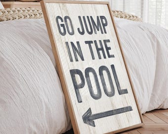 Könnte beinhalten: Ein rechteckiges Holzschild mit hellem Holzrahmen und weißem Hintergrund mit Holzmaserung. Das Schild zeigt den dunkelgrauen Text GO JUMP IN THE POOL in einer fetten serifenlosen Schrift sowie einen nach links zeigenden dunkelgrauen Pfeil.