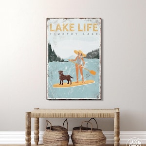 A teal Lake Life paddleboard print of a person and dog gliding across a mountain lake, framed above a bench with baskets