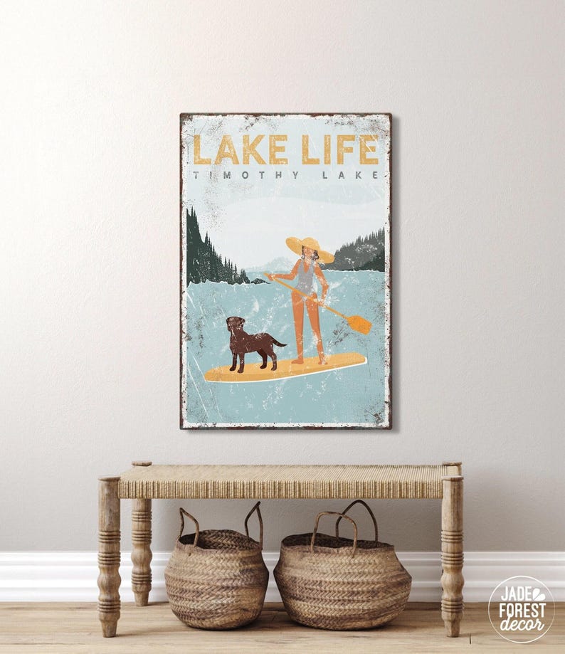 A teal Lake Life paddleboard print of a person and dog gliding across a mountain lake, framed above a bench with baskets