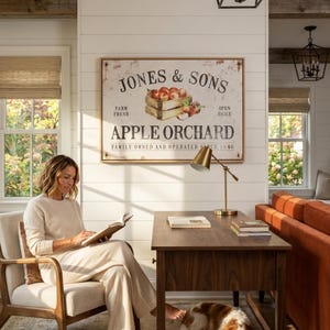 A woman is sitting in a chair, reading a book in a cozy living room with a wooden desk, lamp, and a framed sign for Jones & Sons Apple Orchard.