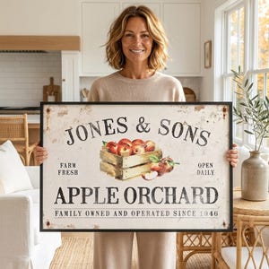 A woman is holding a large sign that says "Jones & Sons Apple Orchard" in a kitchen setting.