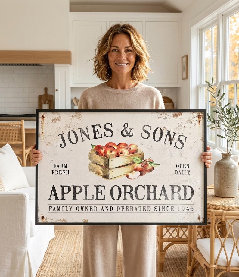 A woman is holding a large sign that says "Jones & Sons Apple Orchard" in a kitchen setting.