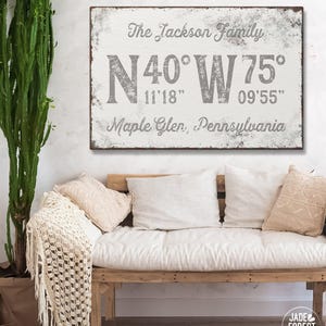 May include: A distressed white sign with gray text reading "The Jackson Family" and coordinates for Maple Glen, Pennsylvania. The sign hangs above a wooden bench with white cushions and pillows. A cactus is in a brown pot to the left.