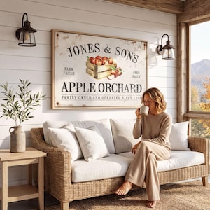 A woman sits on a wicker couch in a cozy room, sipping a beverage and enjoying the view of the mountains outside. Above her, a large framed sign for Jones & Sons Apple Orchard hangs on the wall, featuring a picture of apples and the text "Jones & Sons Farm Fresh Apple Orchard Family Owned and Operated Since 1943".