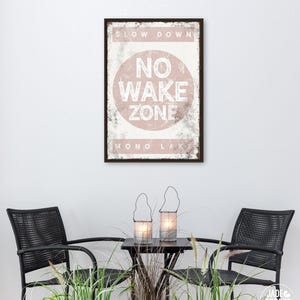 Könnte beinhalten: Ein gerahmtes Schild mit dem Text "SLOW DOWN NO WAKE ZONE MONO LAKE" in Weiß und Rosa. Darunter flankieren zwei schwarze Korbstühle einen kleinen Tisch mit zwei Kerzenlaternen. Hohes grünes Gras und dekorative Pflanzen befinden sich im Vordergrund.