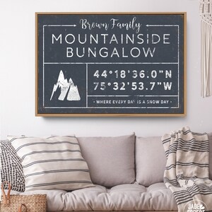 a framed sign with the text "Brown Family MOUNTAINSIDE BUNGALOW" and a mountain icon, mounted on a wall above a couch with a striped blanket draped over it.