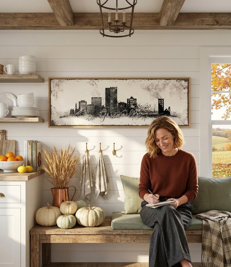 A woman sits on a bench in a cozy kitchen, writing on a notebook. The kitchen features a rustic wooden bench, a large framed cityscape artwork, and various kitchen items such as a bowl, a vase, and a basket of pumpkins.