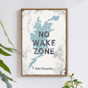 A typographic print reads No Wake Zone with a dusty blue lake silhouette, displayed near a potted plant.