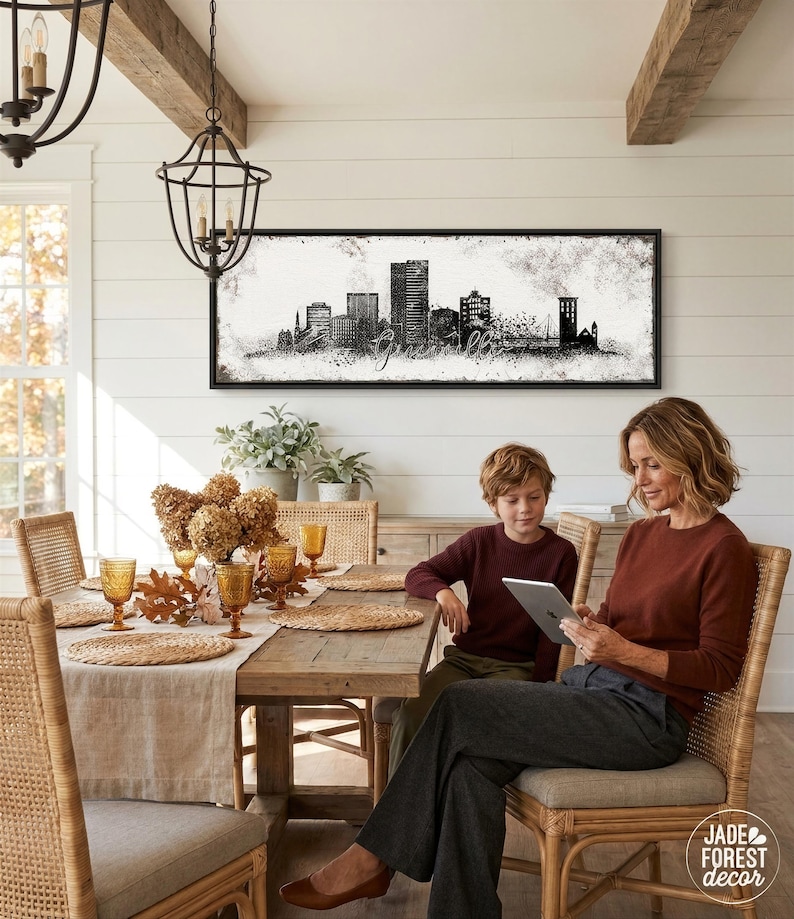 A woman and a young boy are seated at a wooden dining table, with a framed cityscape artwork hanging on the wall above them. The woman is using a tablet, while the boy is looking at the screen. The table is set with various glassware, including wine glasses and cups, and there are potted plants and vases on the table.