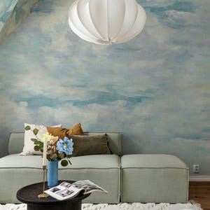 Könnte beinhalten: Ein hellgrünes Modulsofa mit einer weißen Pendelleuchte darüber. Ein dunkelbrauner runder Couchtisch trägt eine blaue Vase mit Blumen und ein aufgeschlagenes Buch. Die Wand zeigt ein blau-weißes Wolkenwandbild und ein weißer Teppich liegt am Boden.
