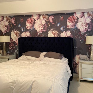 May include: A bedroom featuring a dark blue tufted headboard, a white bed with gray pillows, and two mirrored nightstands with lamps. The wall behind the bed is covered in wallpaper with a large-scale floral pattern in shades of pink, red, and blue.