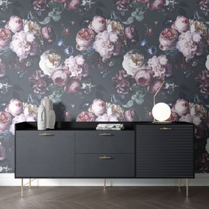 May include: A black sideboard with two drawers, a cabinet door with horizontal grooves, and gold-toned legs. It features gold-toned handles, two gray decorative sculptures, a stack of books, and a round lamp against floral wallpaper.