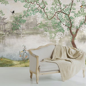 May include: A light beige upholstered settee with a wooden frame and a draped textured throw blanket, positioned against a wall mural featuring a landscape with a pond, a pagoda, a stone bridge, flowering trees, and various birds.
