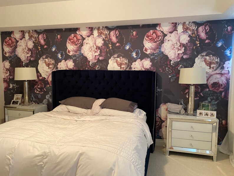 May include: A bedroom featuring a dark blue tufted headboard, a white bed with gray pillows, and two mirrored nightstands with lamps. The wall behind the bed is covered in wallpaper with a large-scale floral pattern in shades of pink, red, and blue.