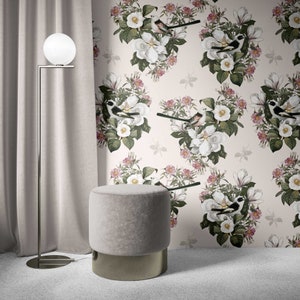 May include: A light grey fabric ottoman with a gold metal base. The ottoman is in front of a wall with a floral wallpaper featuring white flowers, pink flowers, and black birds.