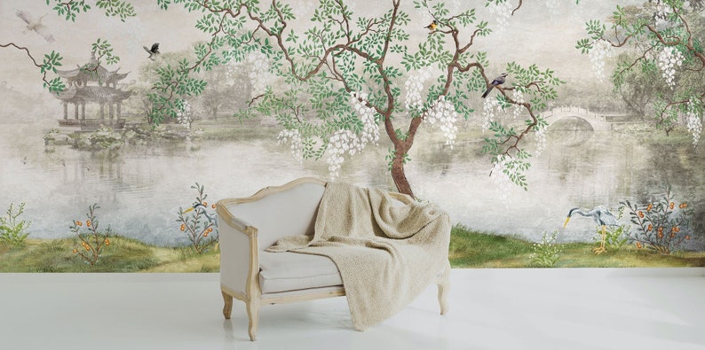 May include: A light beige upholstered settee with a wooden frame and a draped textured throw blanket, positioned against a wall mural featuring a landscape with a pond, a pagoda, a stone bridge, flowering trees, and various birds.