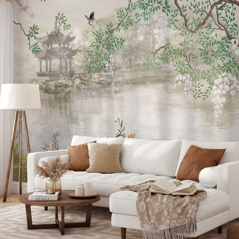 May include: A white tufted sofa with two brown and two beige throw pillows, a wooden coffee table with books and candles, and a floor lamp. The wall features a mural of a pagoda, a pond, and overhanging tree branches with green leaves.