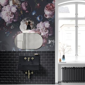 May include: A bathroom interior featuring dark floral wallpaper with large pink, purple, and blue blossoms. A black rectangular wall-mounted sink with gold-toned cross-handle faucets is centered above black subway tiles and below an oval mirror.