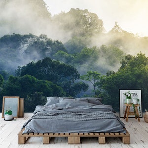 May include: A bedroom featuring a wall mural of a misty green forest, a gray bed with a wooden pallet frame, a framed print with text, a small wooden stool with a potted plant, and a woven basket on a white distressed wooden floor.