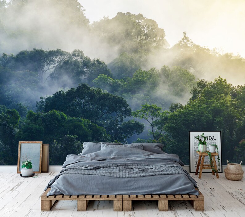 May include: A bedroom featuring a wall mural of a misty green forest, a gray bed with a wooden pallet frame, a framed print with text, a small wooden stool with a potted plant, and a woven basket on a white distressed wooden floor.