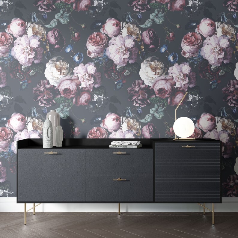 May include: A black sideboard with two drawers, a cabinet door with horizontal grooves, and gold-toned legs. It features gold-toned handles, two gray decorative sculptures, a stack of books, and a round lamp against floral wallpaper.