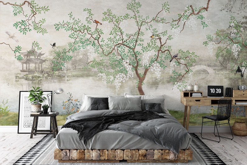 May include: A wall mural featuring a landscape with a large tree, white flowers, green leaves, birds, a pagoda, and a stone bridge. The scene is set behind a bed with grey bedding, a wooden desk with a laptop, and a small side table with a potted plant.
