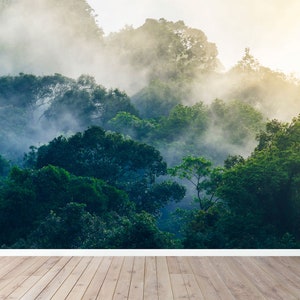 May include: A wall mural featuring a dense, lush green forest canopy shrouded in thick morning mist and fog, with soft golden sunlight filtering through the treetops above, set against a light brown wooden floor with horizontal planks.