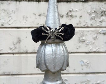 May include: A black velvet hair tie with a silver spider charm adorned with rhinestones. The hair tie is tied around a silver metal finial.