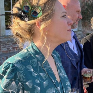 May include: A woman wearing a teal green dress with a white floral pattern. She is wearing a peacock feather hair clip in her hair. She is holding a champagne flute in her right hand.