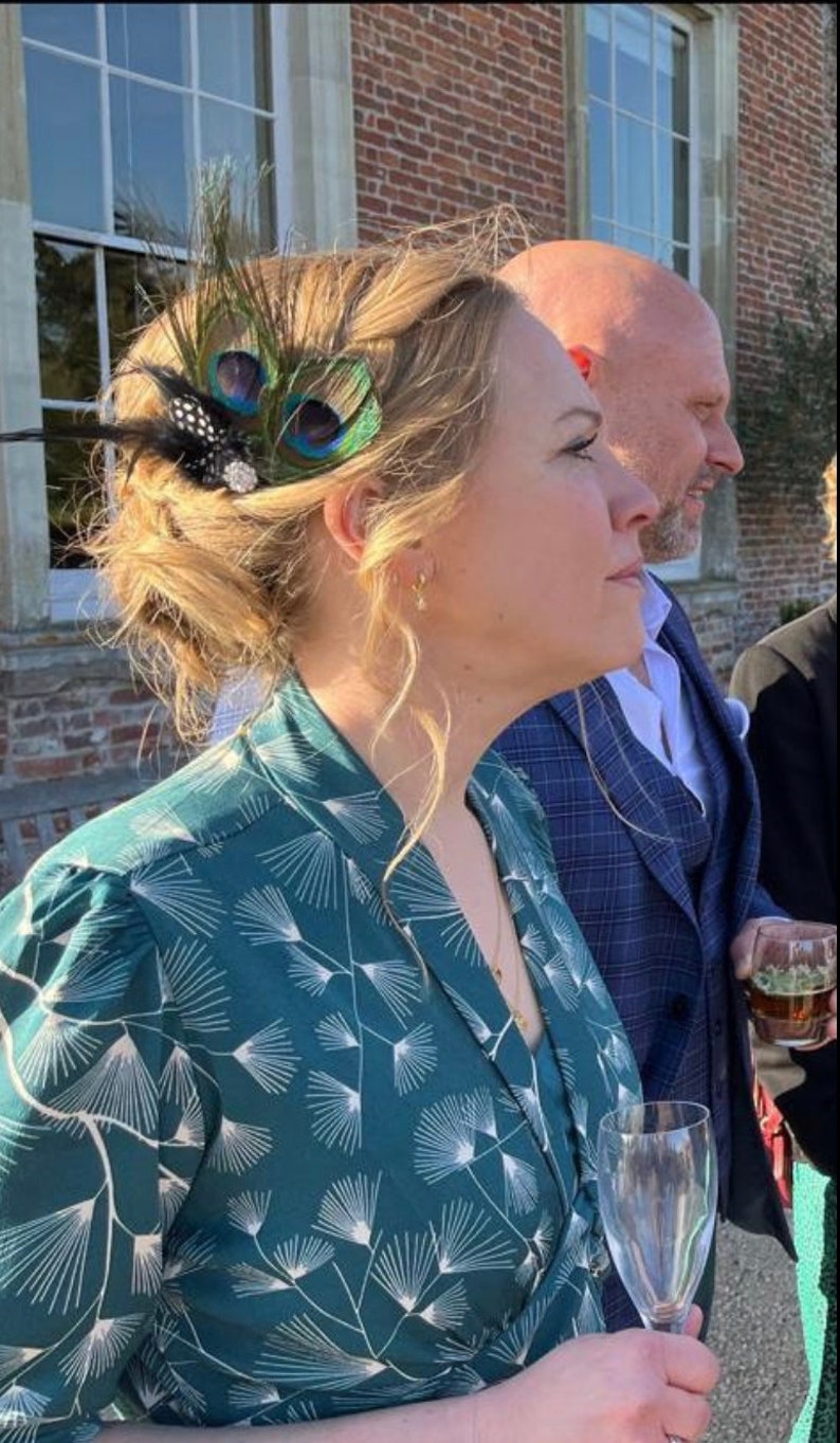 May include: A woman wearing a teal green dress with a white floral pattern. She is wearing a peacock feather hair clip in her hair. She is holding a champagne flute in her right hand.
