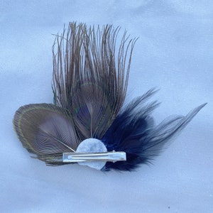 May include: A peacock feather hair clip with a blue and brown feather design. The clip has a silver metal clasp and a white pearl-like embellishment.