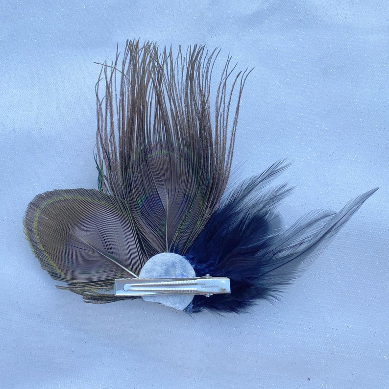 May include: A peacock feather hair clip with a blue and brown feather design. The clip has a silver metal clasp and a white pearl-like embellishment.