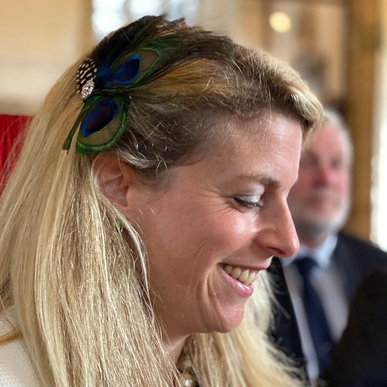 May include: A woman with blonde hair is wearing a peacock feather hair clip. The clip has blue, green, and brown feathers. The woman is smiling and looking down.