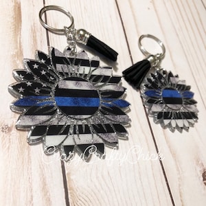 May include: Two silver keychains with black tassels. Each keychain features a blue line American flag sunflower design. The larger sunflower is approximately 3 inches in diameter.
