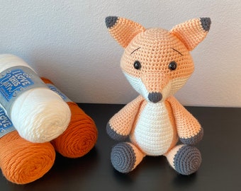 May include: A crocheted fox toy with orange and white fur, black eyes, and gray feet. The toy is sitting on a black surface with two balls of yarn in the background.