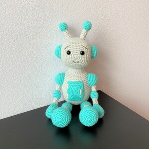 May include: A crocheted robot toy with white and turquoise blue accents. The robot has a smiling face and is sitting on a black surface.