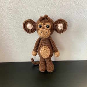 Puede incluir: Un juguete de mono de crochet marrón con orejas rosas y ojos negros. El mono está de pie sobre una superficie negra.