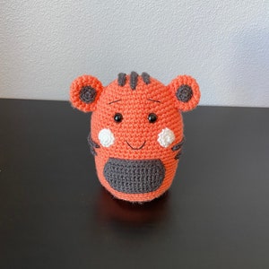 Könnte beinhalten: Ein gehäkeltes orangefarbenes Tiger-Plüschtier mit schwarzen Streifen, schwarzen Ohren und weißen Akzenten um die Augen. Der Tiger hat eine schwarze Nase und ein gesticktes Lächeln.