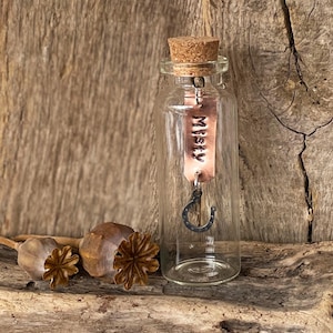 May include: A clear glass bottle with a cork stopper, featuring a copper tag inscribed with "Misty" and a small horseshoe charm. The bottle is set against a rustic wooden background, with dried seed pods nearby.