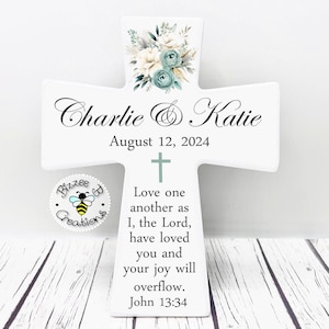 May include: White wooden cross with a floral design in shades of blue and white. The cross is personalized with the names "Charlie & Katie" and the date "August 12, 2024". The cross also features a Bible verse: "Love one another as I, the Lord, have loved you and your joy will overflow. John 13:34".
